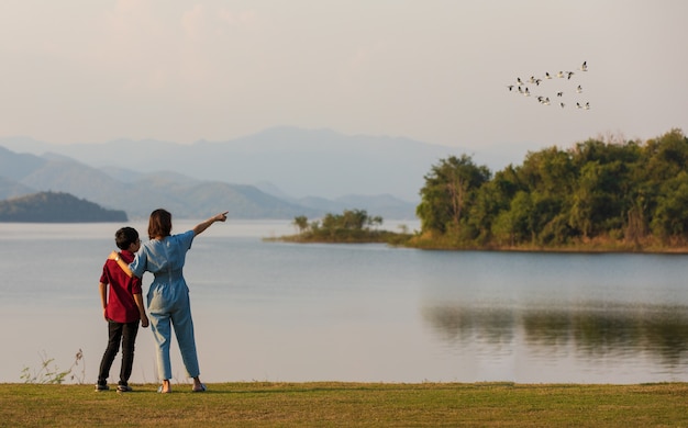 Madre e hijo de pie junto al gran lago y ver la vista de la montaña en el fondo, mamá señalando con el dedo a los pájaros que vuelan en el cielo. Idea para viajes familiares turísticos juntos.