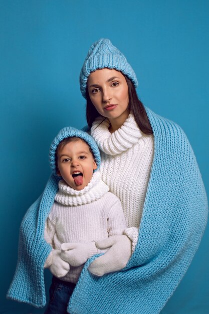 Madre e hijo en suéteres blancos y sombreros azules de pie sobre un azul en el estudio