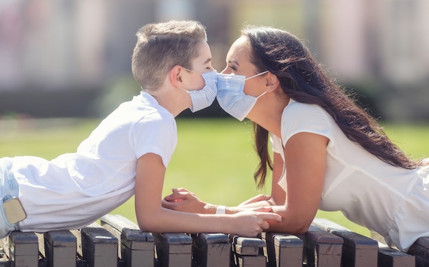 Madre e hijo vestidos con camisetas blancas y máscaras faciales acostados boca abajo en un banco afuera, cara a cara, con la nariz tocándose.