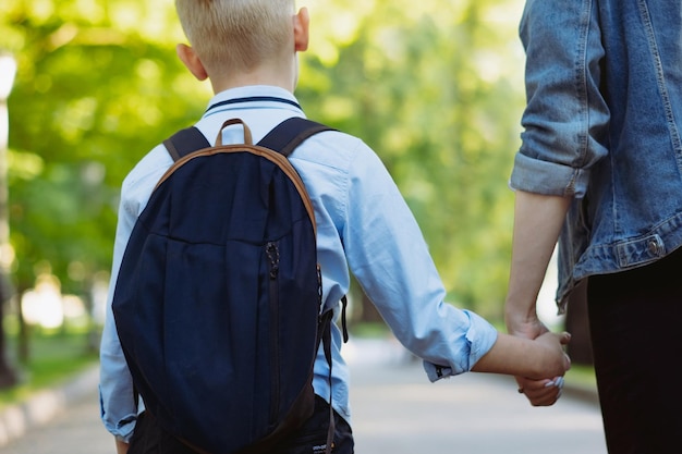 madre e hijo yendo a la escuela tomados de la mano. Muchacho rubio caucásico con pelos atados en cola de caballo mirando a mamá. Imagen con enfoque selectivo