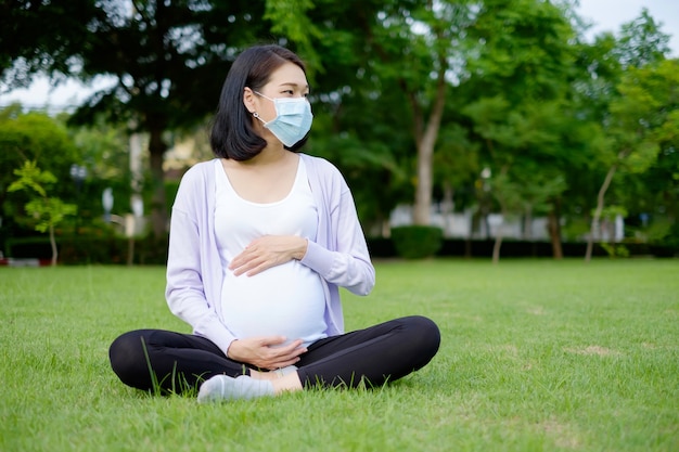 Una madre embarazada con ropa casual de color púrpura y blanco y una máscara se está relajando en el césped.