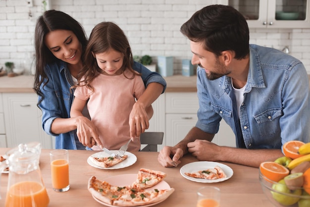 Foto madre enseñando a su hija a usar cubiertos para comer mientras su padre mira en la cocina