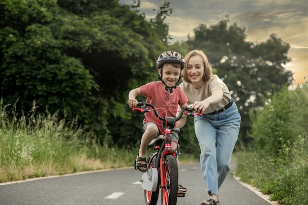 Madre enseñando a su hijo a andar en bicicleta. Chico lindo feliz en casco aprender a andar en bicicleta en el carril bici en verano lloviendo día al atardecer. Fin de semana familiar.