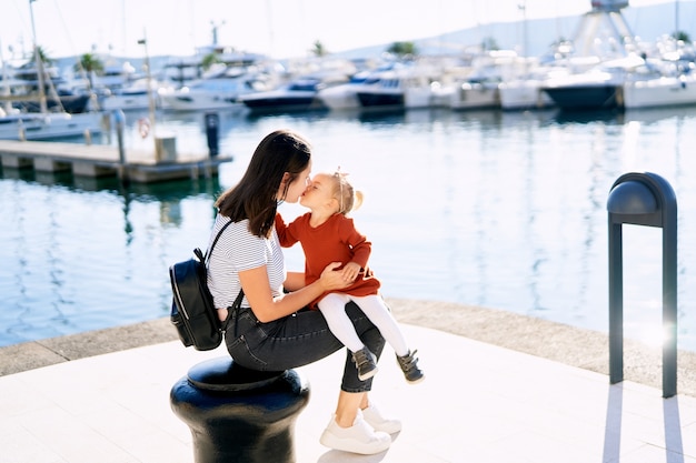 La madre está besando a su bebé mientras está sentada en un muelle junto al mar.