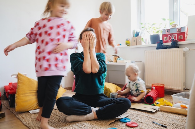 Madre estresada sentada en el suelo en medio de los juguetes mientras los niños traviesos corren alrededor de ella en la habitación Mujer sola agotada con los niños Casa familiar con caos desorden Movimiento borroso para la velocidad vida real