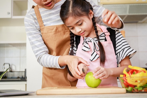 Madre de familia asiática con niña corta la manzana verde para preparar ensalada de verduras en la cocina