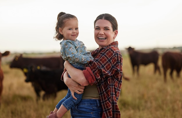 Foto madre de familia y bebé en una granja con vacas en el fondo comiendo pasto sostenibilidad y agricultura feliz madre productora de leche orgánica con su niña y rebaño de ganado afuera en una naturaleza sostenible