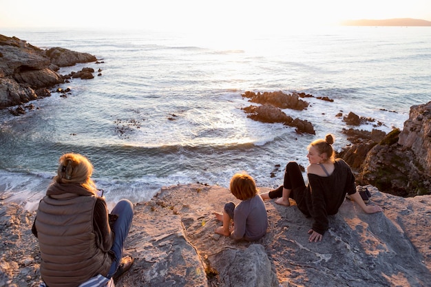 Una madre de familia y dos niños sentados viendo la puesta de sol sobre el océano.