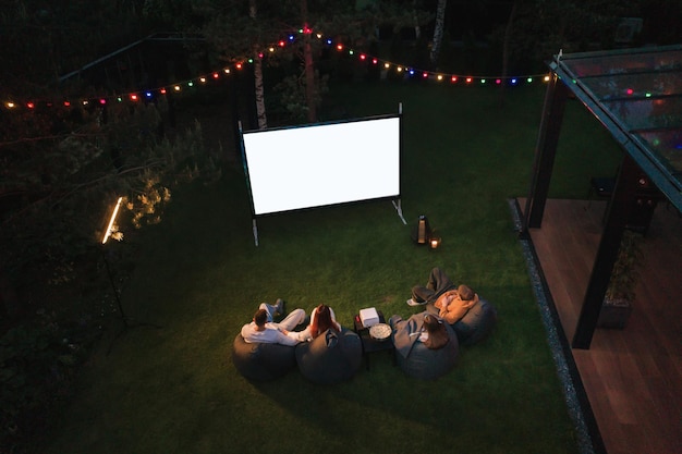Madre de familia, padre e hijos ven un proyector de películas con palomitas de maíz por la noche en el patio
