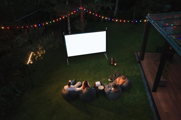 Foto madre de familia, padre e hijos ven un proyector de películas con palomitas de maíz por la noche en el patio