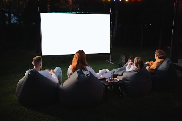 Madre de familia, padre e hijos ven un proyector de películas con palomitas de maíz por la noche en el patio