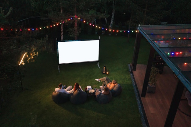 Madre de familia, padre e hijos ven un proyector de películas con palomitas de maíz por la noche en el patio