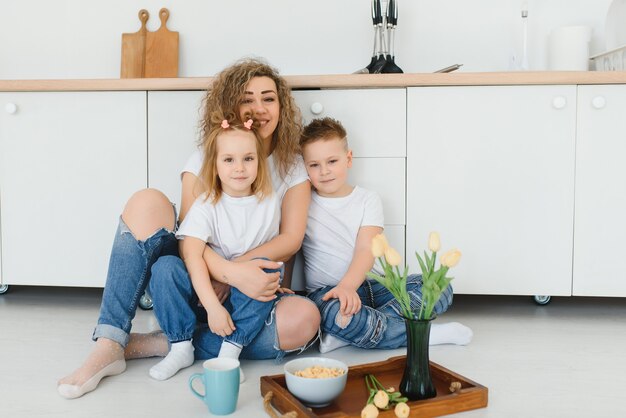 Madre feliz abrazando a su hija e hijo sentados en un piso de madera en la cocina de casa.