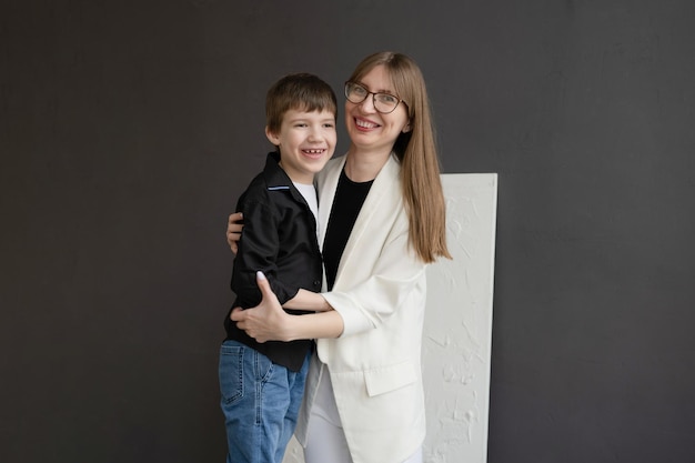 Una madre feliz con anteojos y un hijo en edad preescolar abrazándose y sonriendo en un fondo gris