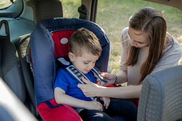 Madre feliz mirando a su hijo en un asiento de bebé. Mujer joven preparando a un niño para un viaje. Concepto de conducción de seguridad.