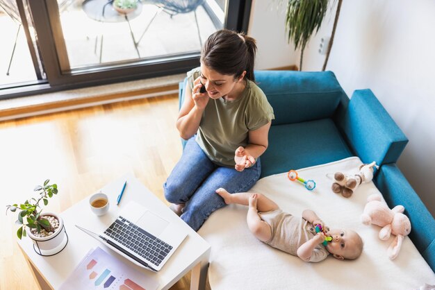 Foto madre feliz ocupada hablando por teléfono mientras cuida a su hija y trabaja en casa.