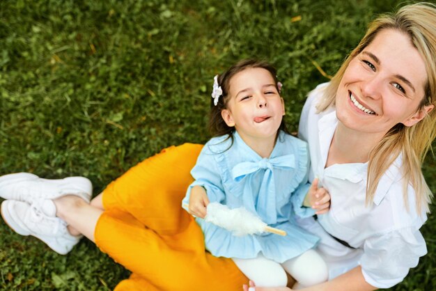 Madre feliz sonriendo y jugando con su hijo disfrutando del tiempo juntos al aire libre Niña alegre comiendo algodón de azúcar con su madre sentada en la hierba verde en el parque de la ciudad Día de la madre