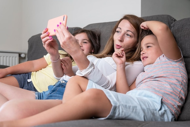 Foto la madre feliz con sus hijos está haciendo una selfie o una videollamada al padre o parientes en un sofá concepto de tecnología paternidad de conexión familiar de nueva generación