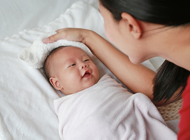 La madre frota el cuerpo seco para la sonrisa del pequeño bebé en la toalla acostado en la cama después del baño