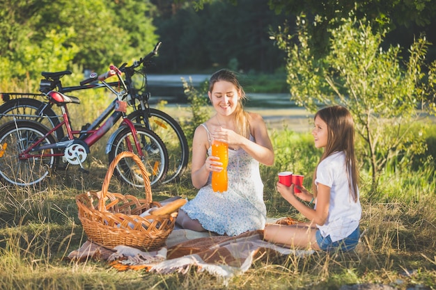 Madre haciendo un picnic junto al río con su hija. Madre vertiendo jugo de naranja en la taza de la hija