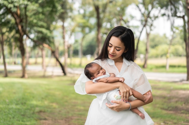 La madre hermosa joven asiática que sostiene a su recién nacido está durmiendo y se siente con amor y se toca suavemente y luego se sienta en la hierba verde en el parque