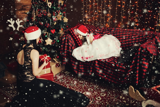 Madre en hermoso vestido negro y sombrero de Santa Claus con presente contra niño dormido en el sofá.