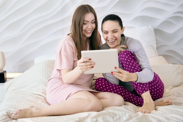 Madre con una hija adulta está viendo videos divertidos en la pantalla de la tableta mientras está sentada en la cama