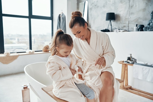 Madre con hija en batas de baño sonriendo y usando crema de manos mientras hace la rutina matutina