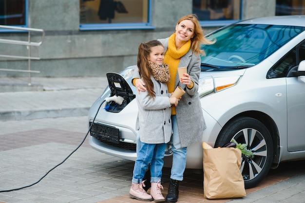 Madre con hija cargando coche eléctrico en la gasolinera eléctrica y hablar por teléfono móvil.