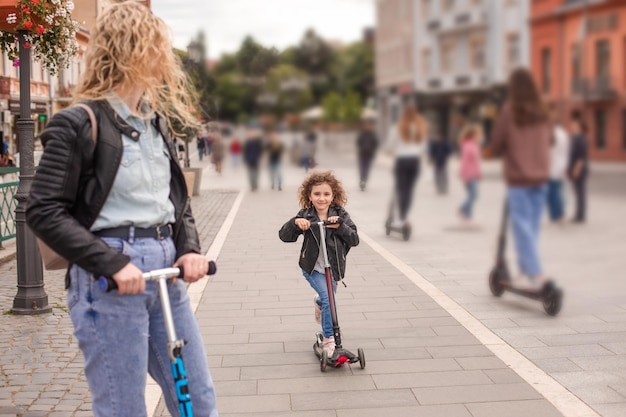 La madre y la hija felices están de pie con scooters en la ciudad