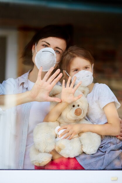 Madre con hija hija en máscara mirando desde la ventana, coronavirus.