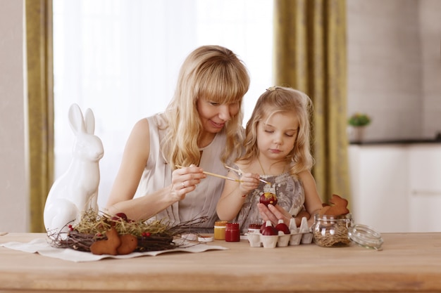 La madre y la hija pintan los huevos de pascua en el cuarto en la tabla del día de fiesta.