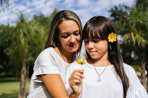 Una madre y una hija sonrientes con una flor, al aire libre.