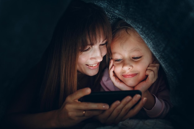 Madre y hija sonrientes mirando el teléfono móvil bajo la manta