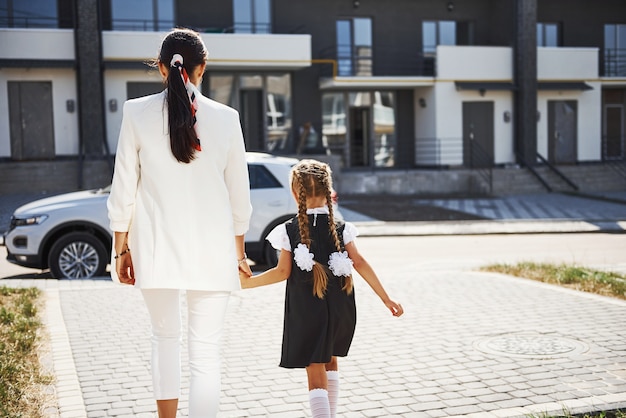 Madre con hija en uniforme escolar al aire libre cerca del coche blanco.