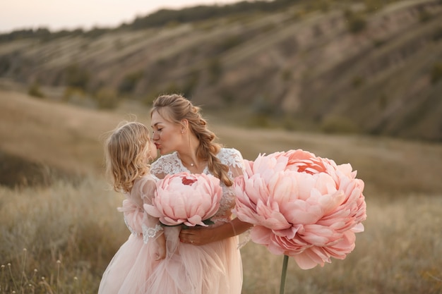 Madre con hija en vestidos de cuento de hadas rosa caminar en la naturaleza. La infancia de la princesita. Grandes flores decorativas de color rosa