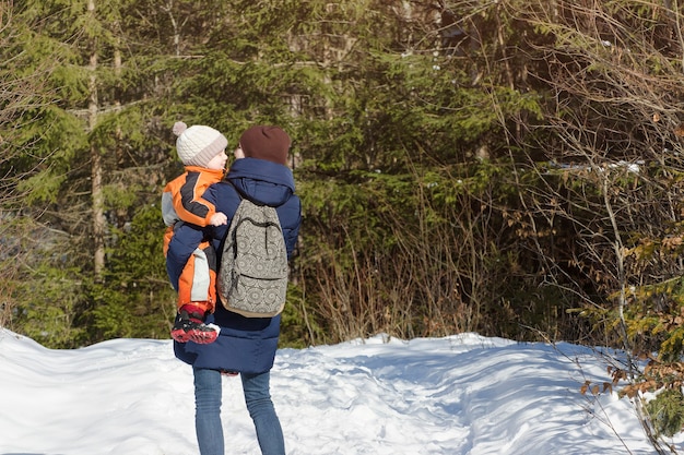 Madre con hijo en brazos y mochila se encuentra en el contexto de bosque de coníferas y camino cubierto de nieve. Vista trasera. Día de invierno, primer plano