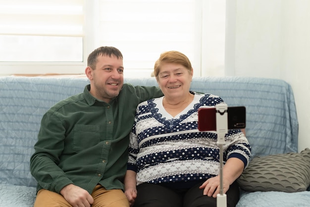 La madre y el hijo mayores sonrientes en casa están sentados en el sofá y hablando por un teléfono móvil