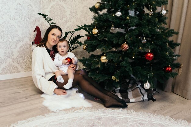 Madre con hijo niño lindo niño juguetón niño pequeño sentado cerca del árbol de Navidad jugando