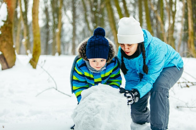 Madre con hijo pequeño haciendo muñeco de nieve rodando gran bola de nieve