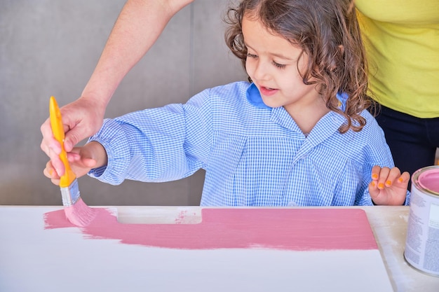 Madre irreconocible ayudando a su hija a pintar un tablero rosa con un pincel