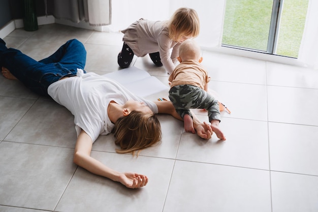 Madre joven y cansada acostada en el suelo relajándose o durmiendo mientras sus hijos dibujan cerca Mujer agotada con niños pequeños en casa Mujer aburrida con niños pequeños Desgaste postparto por maternidad