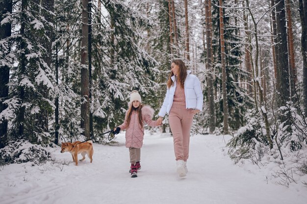 Madre joven de familia feliz y niña linda en ropa de abrigo rosa caminando divirtiéndose con el perro rojo shiba inu en el bosque de invierno frío blanco como la nieve al aire libre. Actividades familiares de vacaciones deportivas.