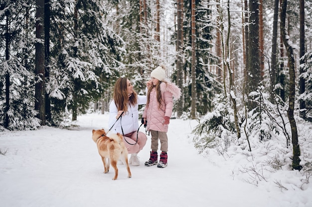 Madre joven de familia feliz y niña linda en ropa de abrigo rosa caminando divirtiéndose con el perro rojo shiba inu en el bosque de invierno frío blanco como la nieve al aire libre