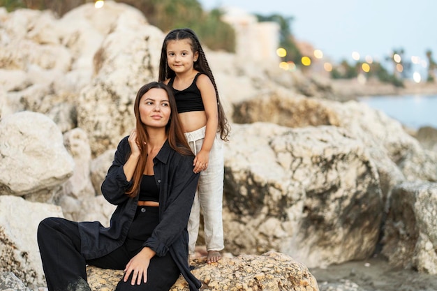 madre joven y hija pequeña posando en las rocas y mirando a la cámara en la playa por la noche