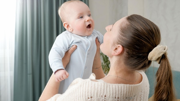 Foto madre joven sosteniendo a su niño sonriente con las manos extendidas frente a una ventana grande