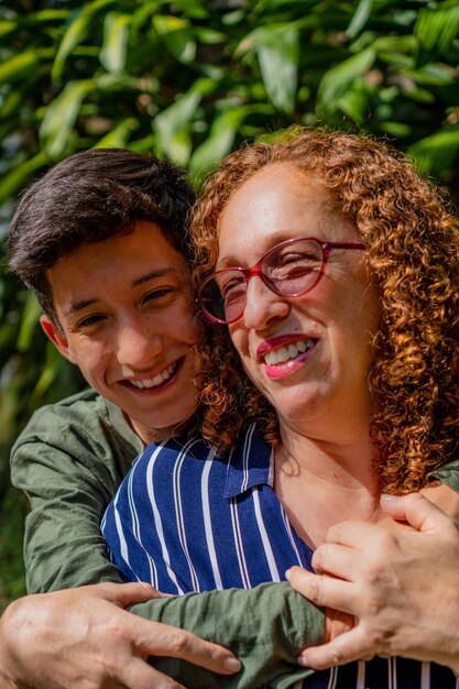 Madre latina de mediana edad con su hijo adolescente abrazándose riéndose en la foto vertical de la naturaleza