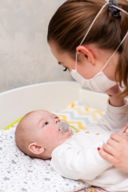 Una madre con una máscara protectora médica cuida a su bebé en su casa en una corontina. Protección profiláctica de coronavirus y covid-19 durante una pandemia.