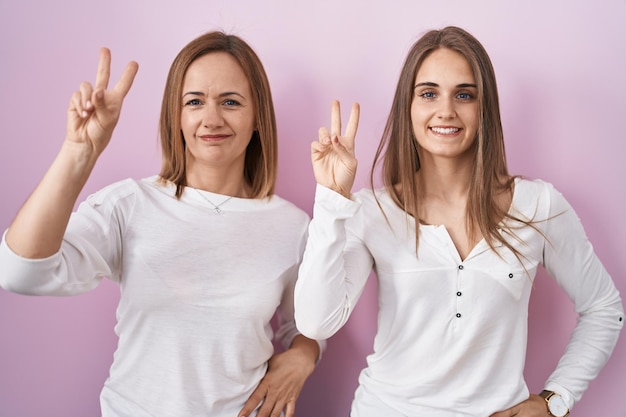 Madre de mediana edad e hija joven paradas sobre un fondo rosado sonriendo mirando a la cámara mostrando los dedos haciendo el signo de la victoria. número dos.