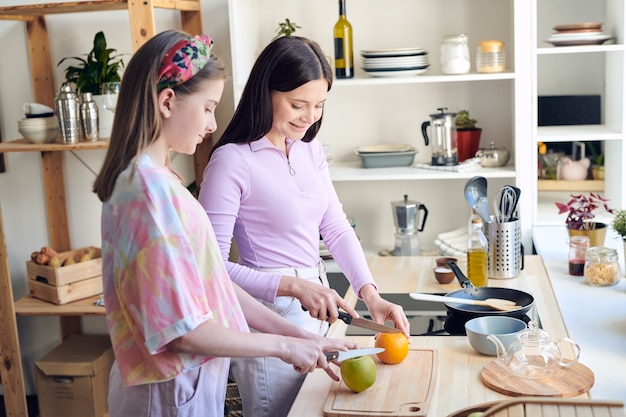 Madre de mediana edad positiva y su hija adolescente de pie en el mostrador de la cocina y cortando frutas mientras preparan el desayuno juntos